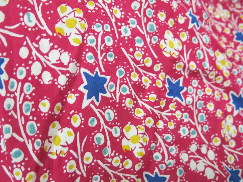 May include: Pink fabric with a white floral pattern. The flowers have yellow centers and are surrounded by blue stars. The fabric is textured and has a slightly wrinkled appearance.