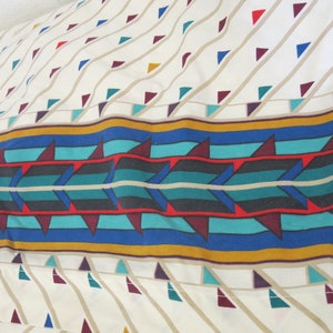 May include: A white fabric with a geometric pattern of red, blue, green, and brown triangles. A band of turquoise, blue, red, and gold stripes runs across the centre of the fabric.
