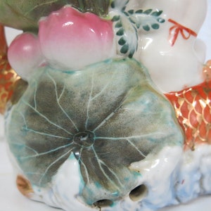May include: A ceramic figurine of a figure riding a koi carp. The carp is orange with gold detailing. The figure is white with a red bow. The figurine includes a lotus flower and leaves.