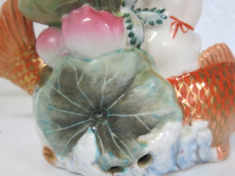 May include: A ceramic figurine of a figure riding a koi carp. The carp is orange with gold detailing. The figure is white with a red bow. The figurine includes a lotus flower and leaves.