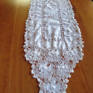 May include: White lace table runner with intricate floral and geometric patterns. The runner features a scalloped edge and openwork details. This decorative textile is suitable for a dining table or dresser.
