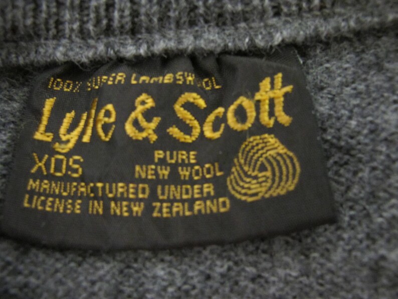 May include: A black label with gold text that reads "100% Super Lambswool Lyle & Scott XOS Pure New Wool Manufactured Under License in New Zealand". The label has a gold yarn ball graphic.