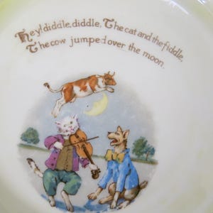 May include: A white ceramic plate with a nursery rhyme illustration. The image features a cow jumping over the moon, a cat playing a violin, and a dog. The plate has the text "Hey! diddle, diddle, The cat and the fiddle, The cow jumped over the moon."