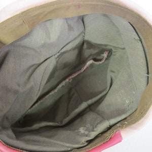 Op de afbeelding: Binnenaanzicht van een roze hoed met een bruine rand en een grijze binnenkant. De hoed heeft een zak en zichtbare stiksels. De rand is een lichtere bruintint en de hoed lijkt vintage te zijn.
