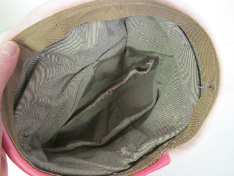 Op de afbeelding: Binnenaanzicht van een roze hoed met een bruine rand en een grijze binnenkant. De hoed heeft een zak en zichtbare stiksels. De rand is een lichtere bruintint en de hoed lijkt vintage te zijn.