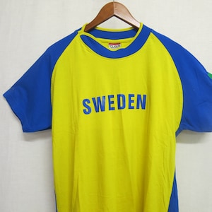 May include: Yellow and blue short-sleeved t-shirt with the word "SWEDEN" printed in blue on the front. The shirt has blue sleeves and a blue collar.
