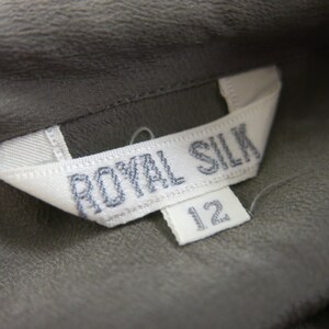 Op de afbeelding: Een wit label met de tekst "ROYAL SILK" in zwarte stiksels. Een kleiner wit label met het nummer "12" is bevestigd aan het grotere label.