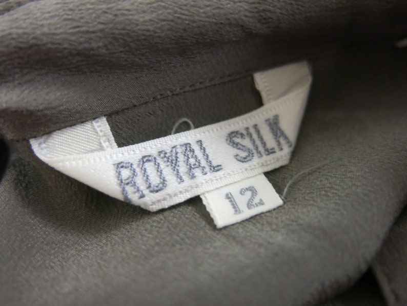 Op de afbeelding: Een wit label met de tekst "ROYAL SILK" in zwarte stiksels. Een kleiner wit label met het nummer "12" is bevestigd aan het grotere label.