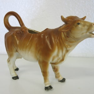 May include: A brown and white ceramic cow creamer with black hooves and a curled tail.