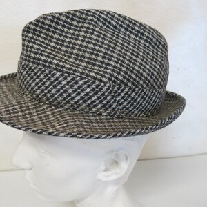 May include: A fedora-style hat with a black and white houndstooth pattern. The hat has a rounded crown and a short brim. The hat is displayed on a white mannequin head.