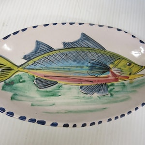May include: An oval ceramic platter featuring a hand-painted fish design in shades of blue, yellow, and green. The fish is centered on a cream-colored background with a decorative border of small, dark blue painted dashes along the outer rim.