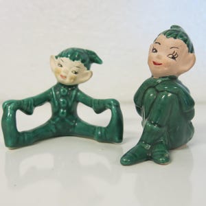May include: Two green ceramic elf figurines. One elf is sitting with its legs crossed and the other elf is sitting with its legs stretched out in front of it.