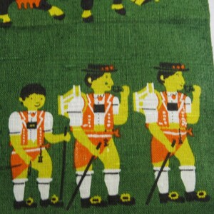 Op de afbeelding: Drie mannen in traditionele Beierse kleding staan op een groene achtergrond. Ze dragen groene hoeden, witte shirts, oranje vesten en oranje broeken. Ze houden wandelstokken vast.
