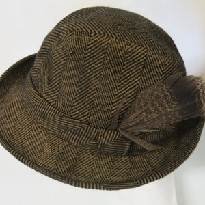 May include: A brown and black herringbone patterned fedora hat with a brown feather accent.