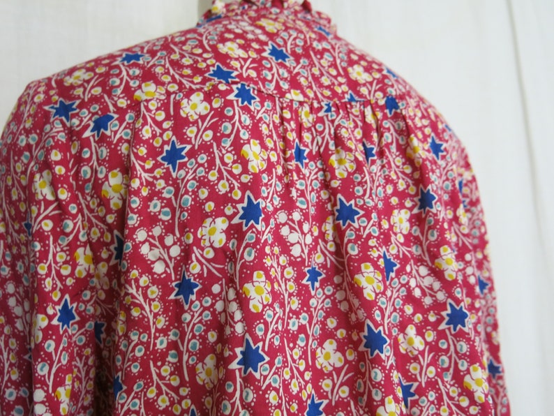 May include: A pink, blue, and white floral print shirt with a star pattern. The shirt has a ruffled collar and a gathered back.