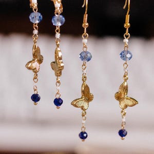 May include: Gold-toned butterfly earrings with blue and white accents. Each earring features a delicate butterfly charm, blue and white beads, and a small, dark blue bead at the bottom. The earrings hang from gold-colored hooks.