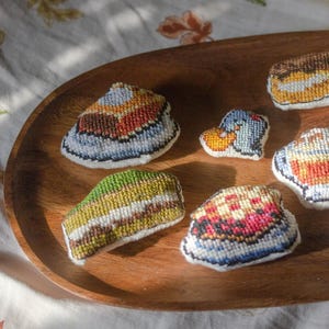Könnte beinhalten: Ein Holztablett mit sieben bestickten Filzkuchen. Die Kuchen sind mit verschiedenen Farben und Mustern verziert, darunter ein blauer und weißer Kuchen, ein grüner und weißer Kuchen, ein roter und weißer Kuchen, ein gelber und weißer Kuchen, ein brauner und weißer Kuchen, ein blauer und weißer Kuchen und ein gelber und weißer Kuchen.