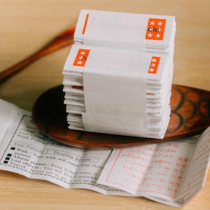 May include: A stack of white fortune cookies with orange star accents and the word "cle" printed on the wrapper. The cookies are on a wooden spoon, with a fortune from one cookie visible.