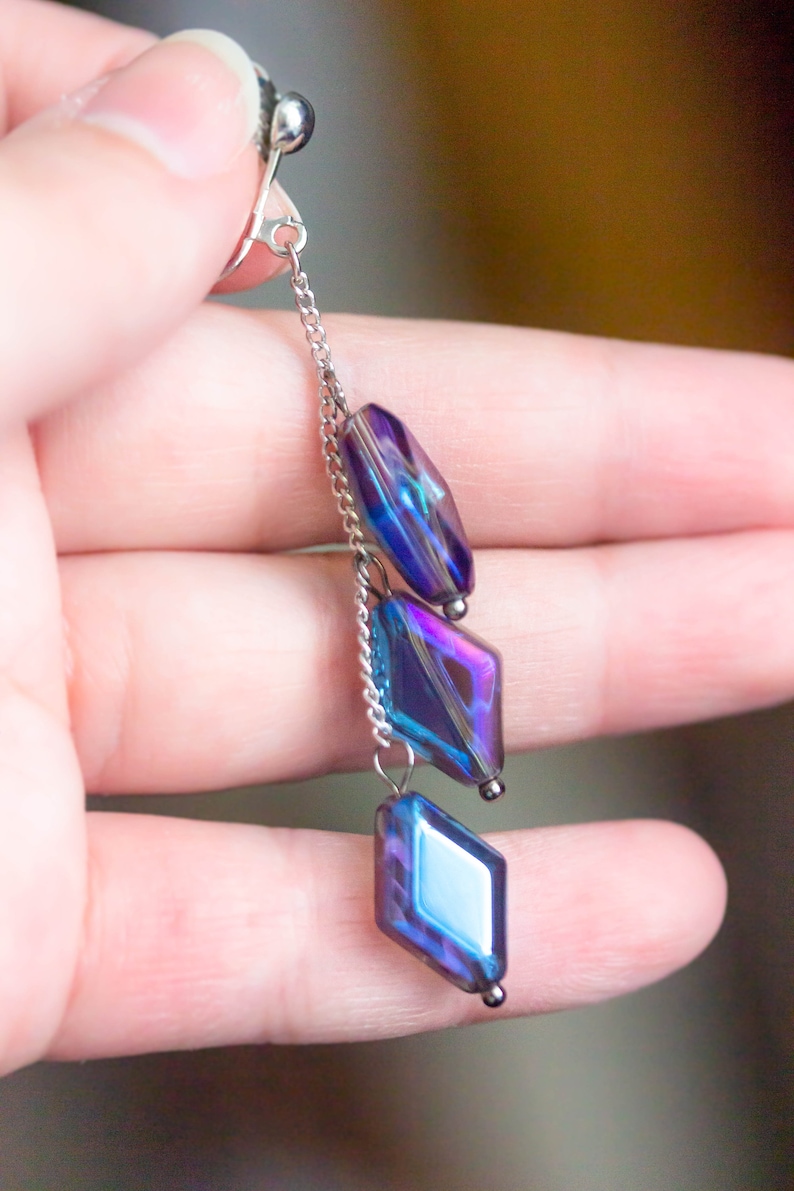 Peut inclure: Une paire de boucles d'oreilles pendantes en verre bleu et violet iridescent avec une fermeture &agrave; clip argent&eacute;e et une cha&icirc;ne.