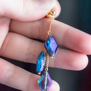 Peut inclure: Une boucle d'oreille pendante dor&eacute;e avec une cha&icirc;ne et trois perles de verre facett&eacute;es bleu et violet iris&eacute;es.