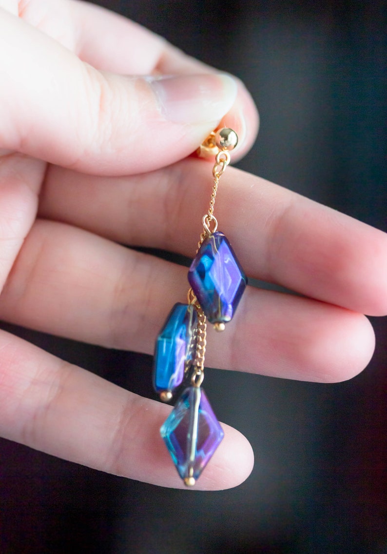 Peut inclure: Une boucle d'oreille pendante dor&eacute;e avec une cha&icirc;ne et trois perles de verre facett&eacute;es bleu et violet iris&eacute;es.