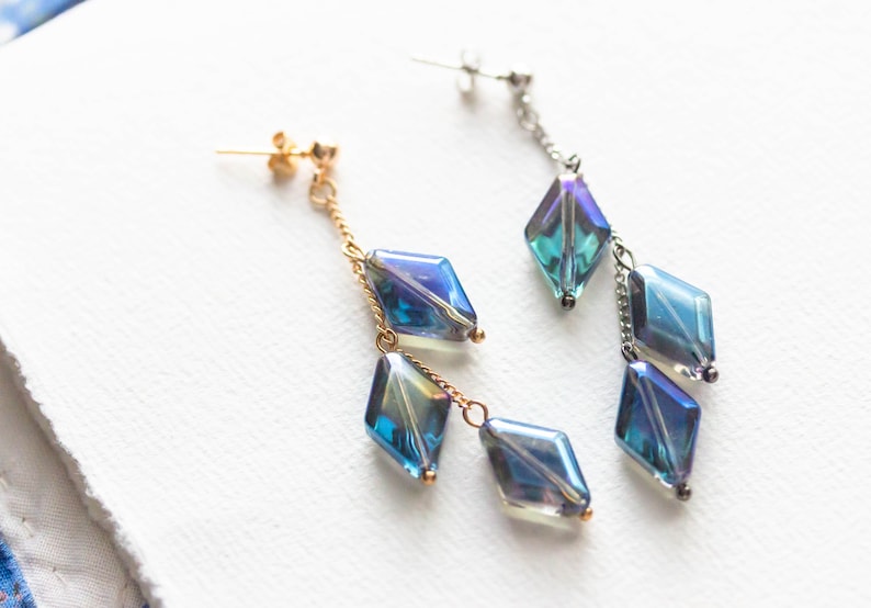 Peut inclure: Une paire de boucles d'oreilles longues et pendantes avec des perles en forme de diamant bleu et vert iris&eacute;. Les boucles d'oreilles ont des cha&icirc;nes dor&eacute;es et argent&eacute;es.