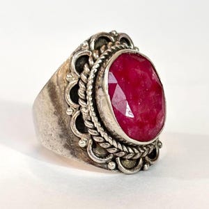 May include: A silver ring featuring a large, oval-cut ruby gemstone. The ruby is a vibrant red color and is set in a decorative silver band with intricate detailing, including a braided rope design around the gemstone and ornate edges.