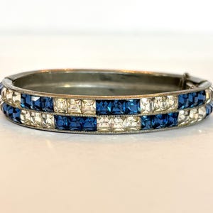 May include: A hinged bangle bracelet featuring two parallel rows of alternating square-cut blue and clear stones set in a silver-toned metal frame. The design displays a repeating checkerboard pattern of deep blue and transparent white crystals.