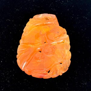 May include: Orange-colored, carved gemstone pendant with intricate floral and leaf designs. The pendant has a smooth, polished surface and is set against a black background. The gemstone is a unique, handcrafted piece.
