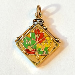 May include: A gold-toned pendant with a colorful maple leaf design. The leaf is divided into sections of green, red, and yellow, with the letters "MWA" visible. The pendant has a diamond shape and a small loop for a chain.