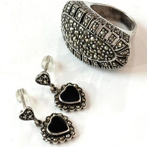 May include: A silver ring and earrings set. The ring has a large, oval face covered in small, round, dark stones. The earrings feature a heart-shaped black stone surrounded by a decorative silver border and a small heart at the top.