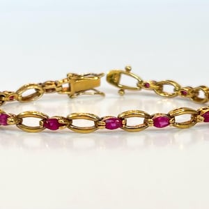 Peut inclure: Un bracelet doré avec des maillons ovales, chacun serti d'une pierre rouge vif. Le bracelet est sécurisé par un fermoir et repose sur une surface blanche, mettant en valeur son design élégant.