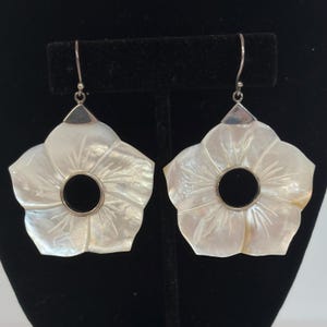 May include: A pair of dangle earrings featuring carved white mother-of-pearl flower petals with a circular cutout in the center. The floral pieces are attached to silver-toned metal hooks with a triangular connector at the top of each earring.