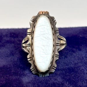 May include: A silver ring featuring an elongated oval white mother-of-pearl inlay set within a scalloped metal bezel. The silver band has a textured, notched edge surrounding the central stone, creating a decorative frame around the inlay.