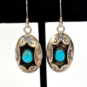 May include: A pair of oval-shaped silver drop earrings featuring a central turquoise stone set within a dark, recessed frame. The silver border is decorated with stamped geometric patterns and each earring hangs from a simple silver hook wire.