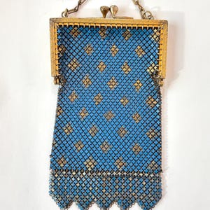 May include: A rectangular mesh purse featuring a bright blue and gold diamond pattern. The bag has a gold-toned metal frame with a dual-knob clasp closure and a matching gold-toned chain strap. The bottom edge of the mesh fabric is scalloped.