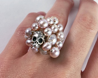 May include: A silver skull ring with a cluster of pink pearls. The ring is on a hand with a white background.