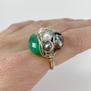 May include: A silver wire wrapped ring featuring a green gemstone, white pearls, and a black pearl.