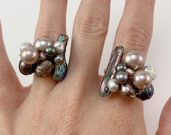 May include: Two rings with a cluster of iridescent pearls and shell pieces. The pearls are in shades of pink, white, and gray. The rings are on a hand.