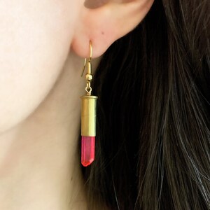 Puede incluir: Pendientes de casquillo de bala dorado con un cristal rosa colgando de la parte inferior.