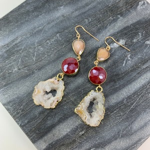 May include: A pair of gold earrings with three gemstone drops. The top drop is a light pink teardrop, the middle drop is a dark red round stone, and the bottom drop is a white agate with a natural crystal formation.