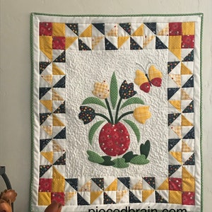 以下が含まれることがあります： 赤、黄色、青のフラワーデザインが施された白いキルト。キルトは黄色、赤、青の三角形のボーダーで縁取られています。キルトは白い壁に掛けられています。キルトの右下隅には「piecedbrain.com」というテキストが印刷されています。