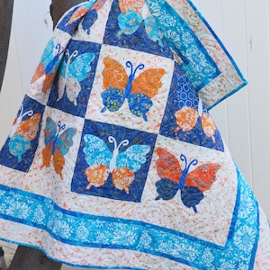 May include: A blue and orange butterfly quilt with a white background. The quilt is made of fabric with a floral pattern and has a blue border.