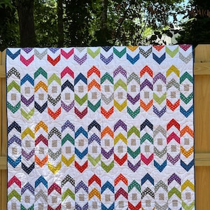 Op de afbeelding: Een kleurrijke quilt met een witte achtergrond en een herhalend patroon van chevronvormen in verschillende kleuren, waaronder blauw, groen, oranje en paars. De quilt hangt aan een houten hek en toont zijn ontwerp en vakmanschap.