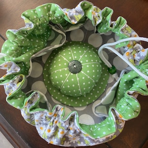 May include: A green and gray fabric pincushion with a drawstring closure. The pincushion is decorated with white polka dots and white daisies.
