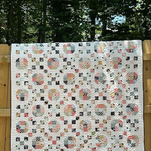 Puede incluir: Un edredón hecho a mano con un fondo blanco y un patrón repetitivo de formas florales y geométricas en colores pastel. El edredón está colgado en una valla de madera al aire libre, mostrando su intrincado diseño y artesanía.