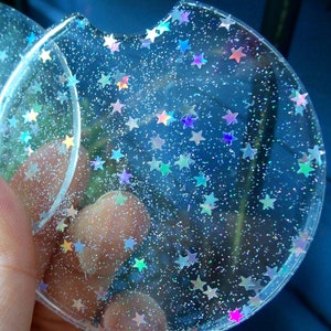 May include: Two clear round coasters with a glittery finish and iridescent stars embedded in the resin. The coasters are translucent and show a blue background through the resin.