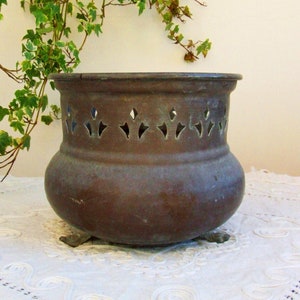 Puede incluir: Una maceta de metal de color bronce envejecido con un cuerpo bulboso y patrones decorativos recortados cerca de la parte superior. La maceta descansa sobre tres pequeños pies y se exhibe sobre una superficie blanca y texturizada. Se ve hiedra verde en el fondo.