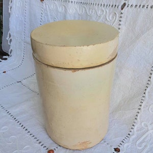 May include: A vintage, off-white ceramic crock with a lid. The crock is cylindrical and has a smooth, slightly textured surface.