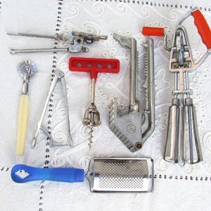 Può includere: Una collezione di utensili da cucina vintage, tra cui un apriscatole, una taglierina per pasta, uno schiaccianoci, un cavatappi, una grattugia e una frusta. Gli utensili sono in metallo con manici rossi, blu e color crema. Disposti su una superficie bianca a motivi.